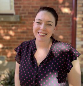Jess is standing in dappled sunlight in front of a red brick wall. She is wearing a black short sleeve top with hot pink spots on it, her hair is pulled back in a ponytail and she is smiling at the camera.