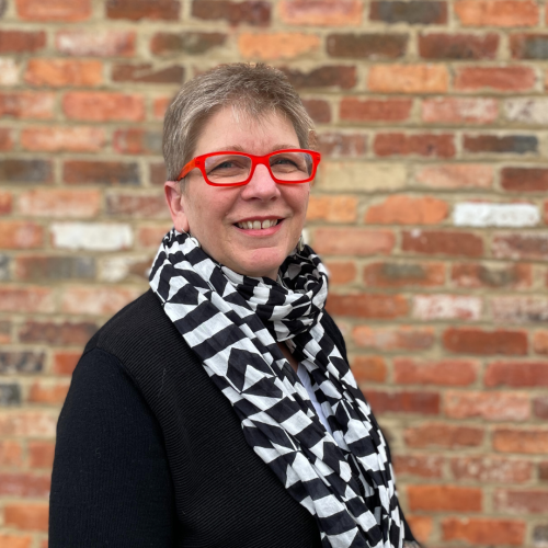 head and shoulders of woman with cropped grey hair, wearing red glasses, a black jacket and black and white striped scarf, standing in front of an old red brick wall