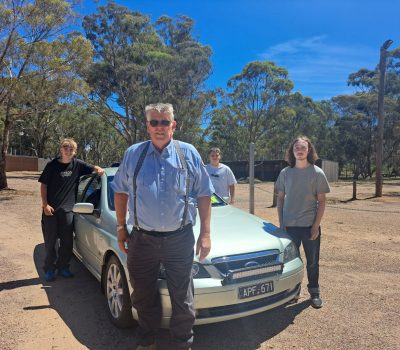 A man in blue shirt and dark pants stands beside a green sedan. Three young learner drivers stand in the background